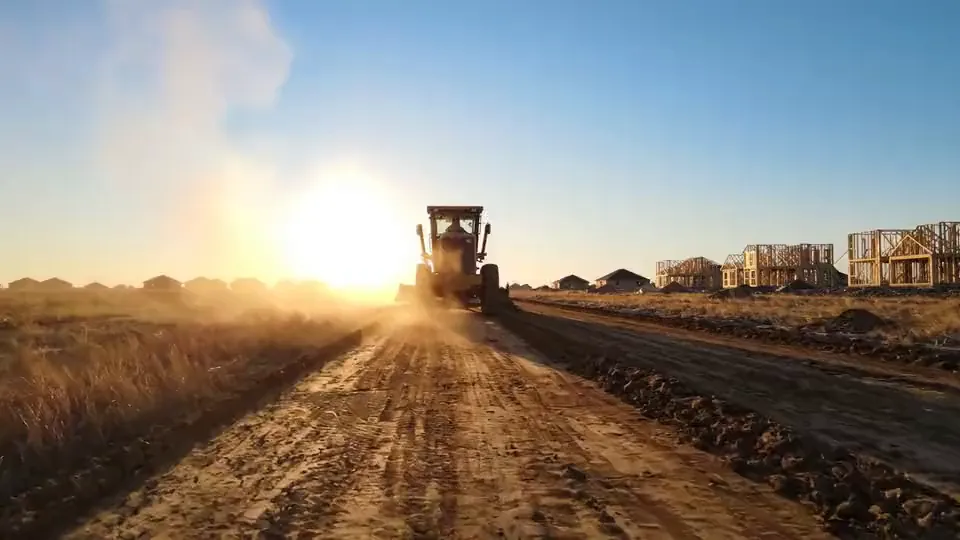 Motor grader shaping a subdivision road with framed houses in the background
